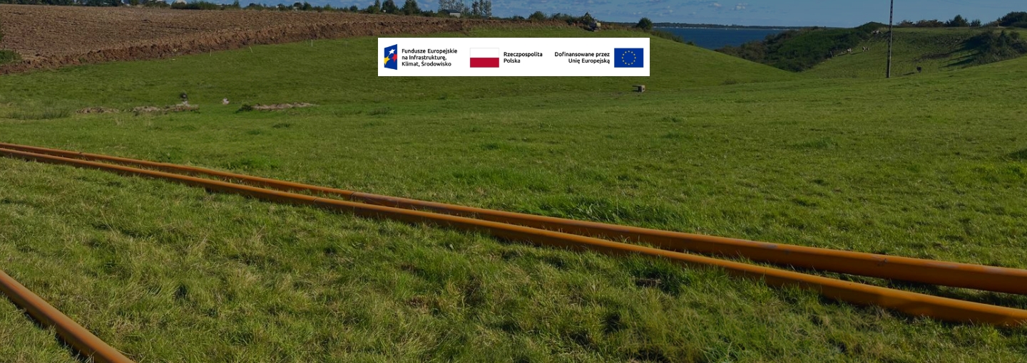 Rury gazowe leżące na trawie, widok na pola, logo programu FEnIKS